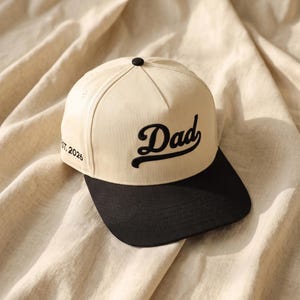 Puede incluir: Una gorra de béisbol color crema con visera negra y un botón negro en la parte superior. El frente presenta la palabra Dad bordada en letra cursiva negra. El costado de la gorra incluye el texto bordado EST. 2026 en letras negras.