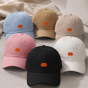May include: A collection of six baseball caps in light blue, tan, beige, pink, black, and white. Each cap features a small, embroidered orange pixelated alien graphic centered on the front panel. The hats have a curved brim and a structured crown.