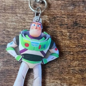 Op de afbeelding: Een Buzz Lightyear sleutelhanger met een wit, groen en paars ruimtepak en een zilverkleurige metalen karabijnhaak. Het figuurtje staat in een staande houding met de handen in de zij en toont gedetailleerde kenmerken van het ruimtepak.
