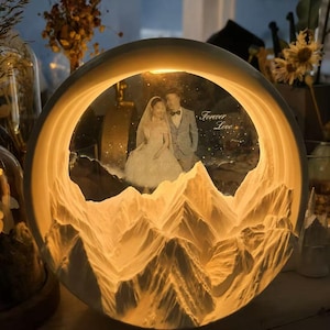 May include: A circular illuminated lamp featuring a 3D white mountain range at the base and a photograph of a couple in wedding attire inside the frame. The text "Forever Love" is printed on the right side of the image, glowing with warm light.