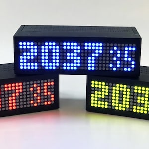 Può includere: Tre orologi digitali rettangolari neri con display a matrice di punti. Ogni orologio mostra l'ora 20:37:35 in diversi colori LED: uno con cifre rosse, uno con cifre blu e uno con cifre giallo-verdi. Gli orologi hanno una trama a griglia.