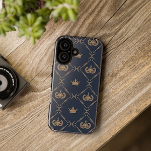 May include: A dark blue smartphone case featuring a repeating pattern of gold-colored crowns and ornate floral flourishes arranged in a diamond grid. The case has a cutout for a dual-lens camera system and a flash on the upper left corner.