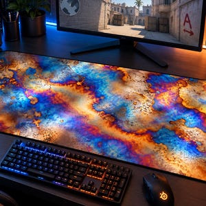 May include: A rectangular desk mat featuring a vibrant, abstract pattern of heat-treated metal textures in shades of deep blue, bright orange, and metallic gold, with a smooth surface and black stitched edges for a clean, finished look.