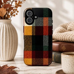 Peut inclure: Une coque de smartphone rectangulaire arborant un motif à carreaux multicolore composé de blocs jaune moutarde, rouge foncé, vert forêt, noir et gris. Le design présente un aspect texturé et inclut une découpe circulaire pour l'objectif.