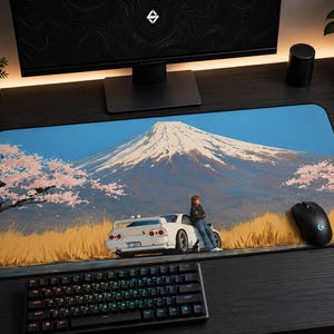 May include: A rectangular desk mat featuring a printed illustration of a white sports car parked in a field of tall yellow grass with a snow-capped mountain in the background and pink cherry blossom trees under a clear blue sky.