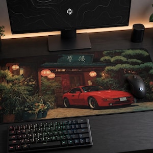 May include: A rectangular desk mat featuring a printed illustration of a red sports car parked in front of a traditional building with glowing lanterns, lush green trees, and Japanese text on a sign above the entrance, with a black computer mouse.