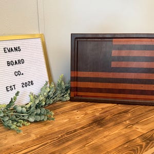 May include: A rectangular wooden cutting board featuring a striped pattern in dark and light brown tones, resembling a flag. Beside it sits a white felt letter board in a gold frame with the text EVANS BOARD CO. EST 2026.