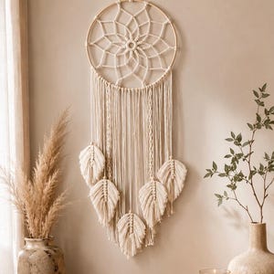 May include: A cream-colored macrame wall hanging featuring a circular wooden hoop with a woven web pattern, long hanging fringe, and five leaf-shaped macrame accents attached to the bottom, suspended by a cord from the top of the hoop.