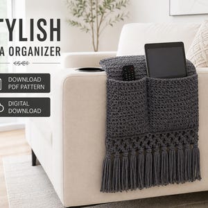 May include: A dark gray crocheted sofa organizer draped over an armrest, featuring two pockets holding a remote and a tablet. The bottom edge has a decorative lattice pattern with long fringe tassels. Text reads STYLISH SOFA ORGANIZER, DOWNLOAD PDF PATTERN, DIGITAL DOWNLOAD.