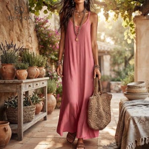 May include: A floor-length dusty pink sleeveless maxi dress with thin spaghetti straps and a scoop neckline. The dress is paired with a large, textured woven straw tote bag and a layered gold-tone pendant necklace.