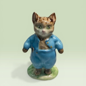 May include: A ceramic figurine of a brown tabby cat standing upright. The cat wears a bright blue jacket with yellow buttons and matching blue trousers. It has green eyes, pointed ears, and stands on a small, circular, light green base.