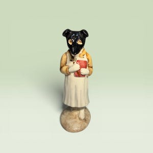 May include: A ceramic figurine of an anthropomorphic dog with a black head, wearing a tan jacket over a white dress. The figure holds a small red rectangular book in its hands and stands on a circular, textured, light brown base.
