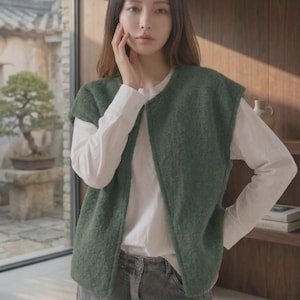 Puede incluir: Un chaleco sin mangas de color verde oscuro con un acabado de punto texturizado, usado sobre una camisa blanca de manga larga y combinado con pantalones vaqueros grises. El chaleco presenta un cuello redondo sencillo, frente abierto y dobladillo recto.