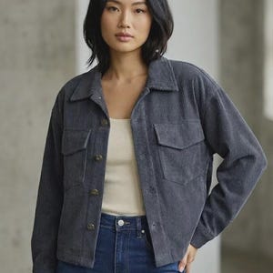 May include: A charcoal grey corduroy jacket featuring a classic collar, long sleeves, and a button-down front. The garment includes two chest pockets with flaps and buttons, showcasing a vertical ribbed texture throughout the fabric construction.