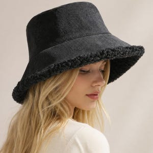 Puede incluir: Un sombrero de pescador negro con una copa estructurada de tela tipo mezclilla y un ala ancha inclinada hacia abajo. La parte inferior y el borde del ala están forrados con un material grueso de piel sintética negra con textura.