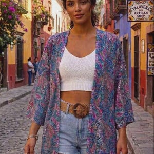 Puede incluir: Un top corto blanco de canalé, pantalones cortos de mezclilla azul claro con dobladillo deshilachado y un cinturón trenzado marrón con una hebilla circular grande. El conjunto incluye un kimono abierto con un estampado de cachemira azul, morado y rosa.