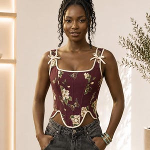 Puede incluir: Un top tipo corsé sin mangas en color burdeos intenso con estampado de flores y mariposas. La prenda presenta un escote cuadrado, ribetes de color crema en los bordes, lazos decorativos en los tirantes y un dobladillo curvo en la cintura.