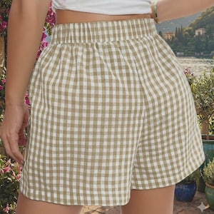 Puede incluir: Pantalones cortos de talle alto con un patrón de cuadros vichy en beige y blanco. Los pantalones cortos tienen una cintura elástica y un ajuste relajado, confeccionados con una tela ligera. El diseño presenta una silueta sencilla y limpia sin bolsillos ni adornos visibles, centrándose en el estampado de cuadros clásico.
