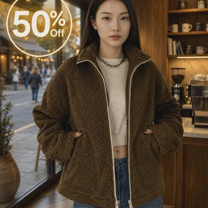 May include: A dark brown, long-sleeved, zip-up fleece jacket with a high collar and white zipper trim. The jacket features a textured, fuzzy material, two side pockets, and is worn over a light-colored top and blue denim jeans. Text reads 50% Off.