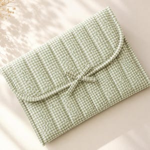 Puede incluir: Un estuche acolchado rectangular con un patrón de cuadros vichy en color verde salvia claro y blanco. El estuche tiene un cierre de solapa redondeado asegurado con una cinta de tela a juego en forma de lazo. El exterior tiene costuras verticales.
