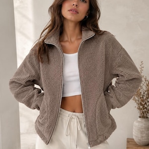 Puede incluir: Una chaqueta de color topo con cremallera y mangas largas, confeccionada en un material de forro polar texturizado y esponjoso. La prenda cuenta con un cuello alto, una cremallera metálica plateada en el centro y dos bolsillos laterales.
