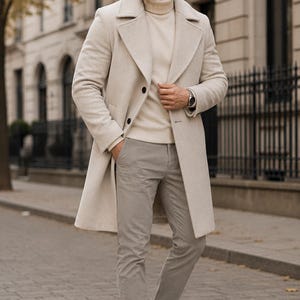 May include: A cream-colored wool-blend overcoat with wide lapels and dark buttons, worn over a matching cream turtleneck sweater and light gray trousers, paired with dark brown suede ankle boots and a silver-toned wristwatch on the wrist.