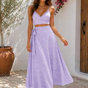 May include: A two-piece set consisting of a lavender crop top with thin spaghetti straps and a V-neckline featuring lace trim, paired with a matching high-waisted midi wrap skirt that has a side tie and a small white speckled pattern throughout.
