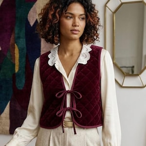 Puede incluir: Un chaleco de terciopelo acolchado color burdeos con dos lazos de cierre frontales, sobre una camisa de manga larga color crema con cuello de encaje festoneado blanco y pantalones claros con un cinturón de tono dorado.