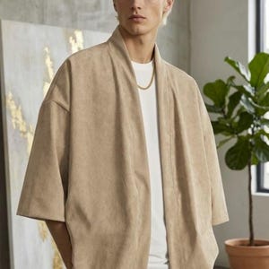 Puede incluir: Una chaqueta beige estilo kimono abierta por delante, con mangas anchas de tres cuartos y una textura suave similar al ante. La prenda se lleva sobre una camiseta blanca lisa de cuello redondo y se combina con pantalones beige.