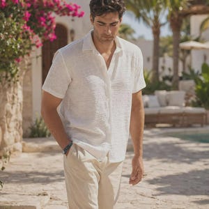 Puede incluir: Una camisa blanca de manga corta con acabado de tela texturizada y cuello clásico, combinada con pantalones de color beige claro. La camisa presenta un corte relajado con dobladillo recto y detalles de botones visibles en la parte frontal.