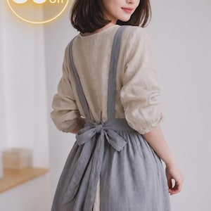 May include: A beige long-sleeved dress featuring a grey apron-style overlay with wide shoulder straps and a large bow tied at the waist. The garment includes a circular graphic in the upper left corner displaying the text 50% Off in white.