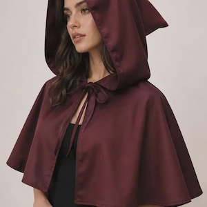 Op de afbeelding: Een diepbordeauxrode cape met capuchon en een gladde, satijnachtige afwerking. Het kledingstuk is voorzien van een puntige capuchon en een bijpassende stoffen striksluiting bij de halslijn, die over de schouders valt voor een gestructureerd effect.
