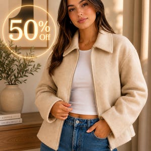 May include: A beige zip-up jacket with a collared neckline worn over a white top and blue denim jeans. The jacket features long sleeves and a straight hem. A glowing circular graphic with the text 50% Off is overlaid on the upper left side.