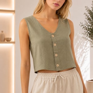 Puede incluir: Un top corto sin mangas de color verde oliva con escote en V y una tapeta frontal con cuatro botones de madera clara. El top se combina con pantalones beige de cintura alta que tienen una cinturilla elástica y un cordón ajustable.