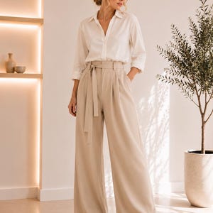 May include: A long-sleeved white button-down shirt with a collar paired with high-waisted beige wide-leg trousers. The trousers feature a matching fabric belt with a tie closure at the waist, front pleats, and a full-length, loose-fitting silhouette.