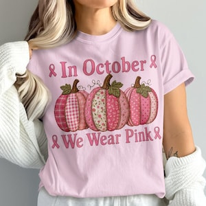 May include: A light pink t-shirt featuring three pumpkins with various pink patterned designs, including plaid and floral prints. The text "In October We Wear Pink" is printed in pink lettering, accompanied by pink awareness ribbons on each side.