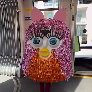 Può includere: Un costume a figura intera che ricorda una creatura con lunghe frange metalliche rosa nella metà superiore e frange metalliche arancioni nella metà inferiore. Presenta grandi occhi bianchi con lunghe ciglia nere, un becco giallo e due alte orecchie rosa.
