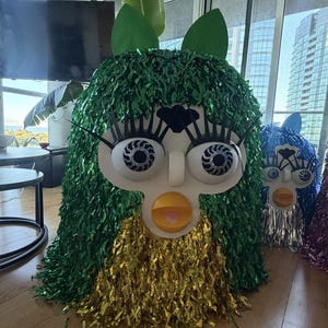 May include: A large, creature-like figure covered in green and gold metallic fringe. It features a white faceplate, large eyes with black spiral pupils, long black eyelashes, a small black nose, and a yellow beak with a pink tongue visible.