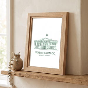 May include: A framed print featuring a green line drawing of the White House in Washington DC, with the text "WASHINGTON DC" and the coordinates "38.9072° N, 77.0369° W" printed below the illustration, all encased in a light-toned wooden frame.