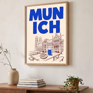 Puede incluir: Una lámina enmarcada con la palabra MUNICH en letras azules llamativas sobre un dibujo lineal de un paisaje urbano, una mesa de madera, un pretzel y una jarra de cerveza azul. La ilustración tiene un fondo crema con líneas rojo oscuro.