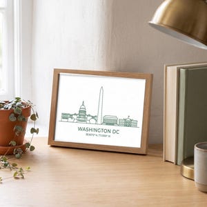 May include: A rectangular wooden frame containing a white print with a dark green line art illustration of the Washington DC skyline. The text reads WASHINGTON DC followed by the coordinates 38.9072° N, 77.0369° W in a simple font.
