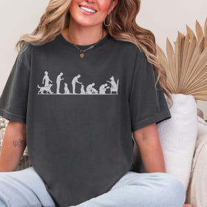May include: A charcoal gray short-sleeve t-shirt featuring a white graphic print that depicts the evolution of a human and a dog, showing the dog eventually sitting on a chair wearing a crown while the human kneels before it.