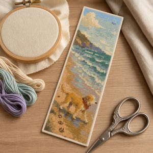 May include: A rectangular cross-stitch bookmark featuring a coastal scene with a golden dog walking on the sand near turquoise ocean waves, a blue sky with clouds, and a pair of ornate silver scissors placed next to the finished embroidery piece.