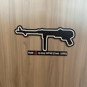 Könnte beinhalten: Ein schwarzes Wand-Schild in Form einer MP40-Maschinenpistole mit weißer Umrandung. Unter der Waffen-Silhouette befindet sich ein rechteckiges schwarzes Schild mit weißem Text: Hold [Quadrat] to buy MP40 [Cost: 1000].