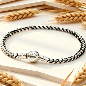 Può includere: Un bracciale circolare caratterizzato da un cordino nero intrecciato con maglie in metallo argentato lucido. Il bracciale include una chiusura sferica in argento con inciso il nome PANDORA, posizionata centralmente per chiudere il gioiello.