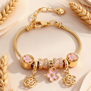 Puede incluir: Una pulsera de dijes con cadena de serpiente dorada que presenta dos cuentas redondas con flores de esmalte rosa, dos cuentas cilíndricas con incrustaciones de patrones rosas y tres dijes colgantes, incluyendo dos formas florales caladas.
