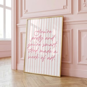 May include: A rectangular framed wall art piece featuring a light pink and white vertical striped background. The text in a dark pink cursive font reads: You're pretty and you're smart. God made a work of art. The frame is a thin metallic gold.