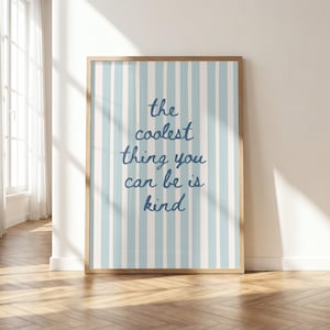 Può includere: Una stampa da parete rettangolare in una cornice di legno chiaro con strisce verticali azzurre e bianche. Al centro, il testo scritto a mano in blu scuro recita: the coolest thing you can be is kind, centrato sullo sfondo a righe.