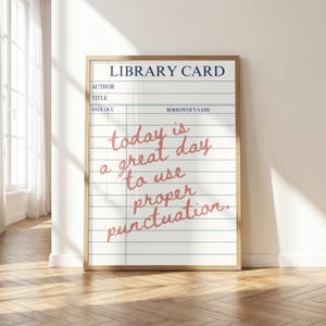 May include: A framed wall art print featuring a white library card template with blue horizontal lines and a vertical margin. The text "LIBRARY CARD" appears in dark blue at the top, while the pink cursive text reads "today is a great day to use proper punctuation."