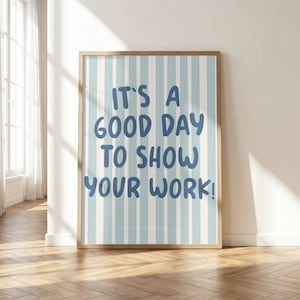 May include: A rectangular wall art print in a light wooden frame featuring vertical light blue and white stripes. The center of the print displays the dark blue text "IT'S A GOOD DAY TO SHOW YOUR WORK!" written in a rounded, casual, hand-lettered font style.